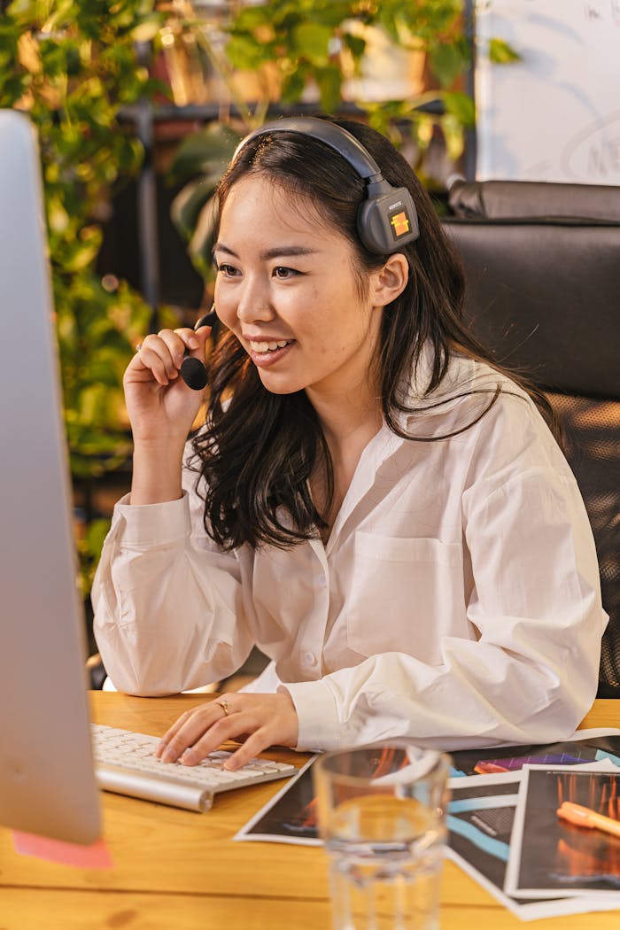 Asian woman in white shirt working on a computer with headset, indoors, smiling.